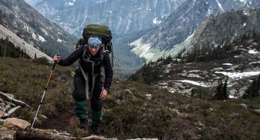 a woman wearing a backpack hikes across rocky terrain with mountains surrounding her